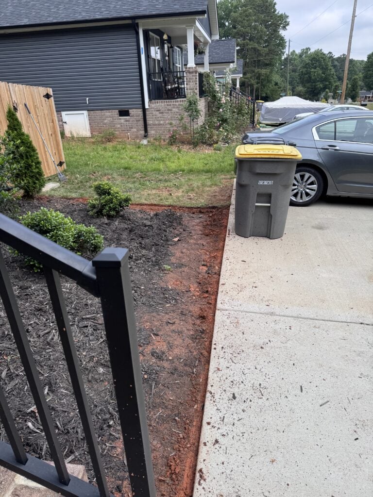 A freshly cut spade edge separates a foundation bed from a concrete driveway, showing exposed Carolina clay and the vertical bed-side face that holds mulch in place.